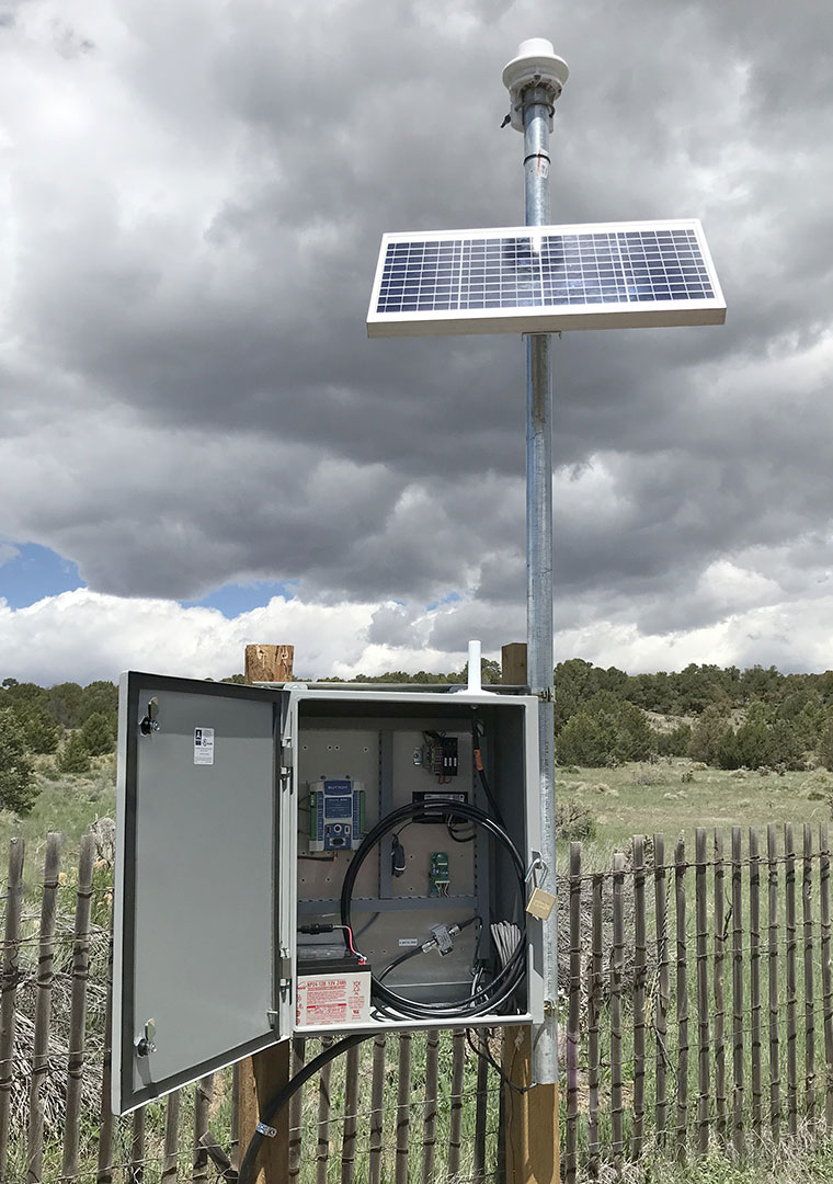 Open enclosure with datalogger and battery. External antenna, solar panel, and Lufft WS100 precipitation sensor with hill and vegetation in the background.