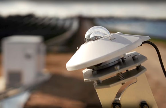 A Kipp & Zonen pyranometer in the foreground is mounted to a bracket. A blurred outdoor environment is shown in the background.